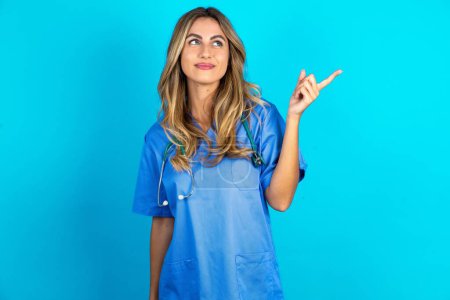 Téléchargez les photos : Belle médecin femme debout sur fond bleu studio regardant caméra indiquant doigt vide espace ventes - en image libre de droit