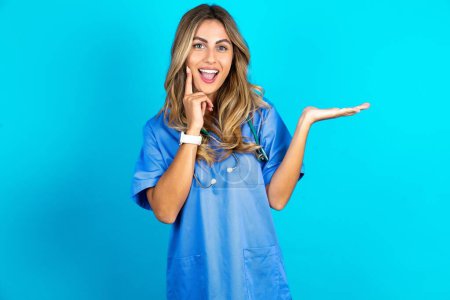 Téléchargez les photos : Funny belle médecin femme debout sur fond bleu studio tenir palme ouverte nouveau produit grande proposition - en image libre de droit
