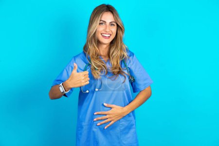 Foto de Hermosa médico mujer de pie sobre azul estudio fondo feliz sonrisa positiva manos en el vientre mostrar pulgar hacia arriba bien saludable - Imagen libre de derechos