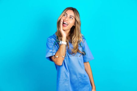 Téléchargez les photos : Belle médecin femme debout sur fond bleu studio entendre incroyable nouvelles privées impressionné partager cri - en image libre de droit