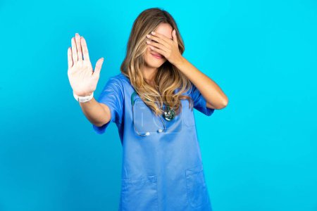 Photo for Beautiful doctor woman standing over blue studio background covers eyes with palm and doing stop gesture, tries to hide from everybody. - Royalty Free Image