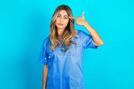 Photo for Unhappy beautiful doctor woman standing over blue studio background makes suicide gesture and imitates gun with hand, curves lips keeps two fingers on temple, shoots, being tired of everything, - Royalty Free Image
