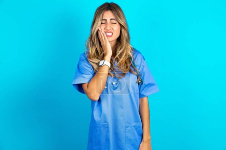 Photo for Beautiful doctor woman standing over blue studio background touching mouth with hand with painful expression because of toothache or dental illness on teeth. - Royalty Free Image