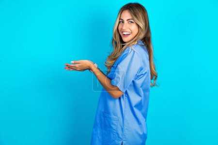 Téléchargez les photos : Belle médecin femme debout sur fond bleu studio pointant de côté avec les mains ouvertes paumes montrant l'espace de copie, présentant la publicité souriant heureux heureux - en image libre de droit