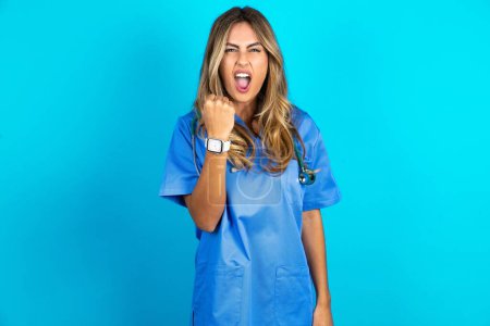 Photo for Beautiful doctor woman standing over blue studio background angry and mad raising fist frustrated and furious while shouting with anger. Rage and aggressive concept. - Royalty Free Image