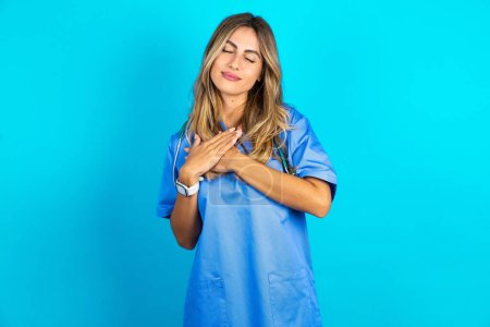 Photo for Beautiful doctor woman standing over blue studio background smiling with hands on chest with closed eyes and grateful gesture on face. Health concept. - Royalty Free Image