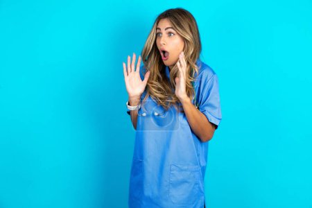 Photo for Beautiful doctor woman standing over blue studio background shouts loud, keeps eyes opened and hands tense. - Royalty Free Image