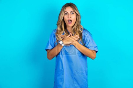 Photo for Scared beautiful doctor woman standing over blue studio background looks with frightened expression, keeps hands on chest, being puzzled to notice something strange, People, hush reaction and emotions. - Royalty Free Image