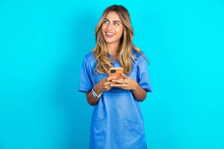 Foto de Hermosa mujer doctora de pie sobre fondo de estudio azul sosteniendo un teléfono inteligente y mirando hacia los lados en blanco copyspace. - Imagen libre de derechos
