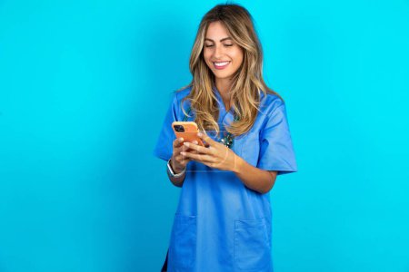 Foto de Hermosa mujer doctora de pie sobre fondo azul estudio utilizando el teléfono móvil chat tiempo libre . - Imagen libre de derechos
