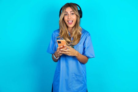 Téléchargez les photos : Magnifique médecin excité femme debout sur fond bleu studio tenant smartphone et regardant étonné de la caméra après avoir reçu de bonnes nouvelles. - en image libre de droit