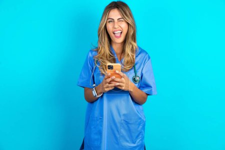Téléchargez les photos : Excité belle médecin femme debout sur fond bleu studio clin d'oeil et tenir l'oeil utilisation de téléphone intelligent lire les nouvelles des réseaux sociaux - en image libre de droit