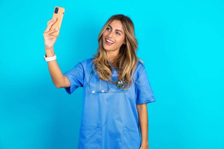Foto de Hermosa doctora de pie sobre fondo azul estudio sonriendo y tomando una selfie listo para publicarlo en sus redes sociales. - Imagen libre de derechos