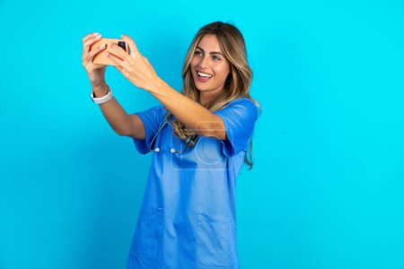Téléchargez les photos : Belle médecin femme debout sur fond bleu studio prendre un selfie pour le poster sur les médias sociaux ou avoir un appel vidéo avec des amis. - en image libre de droit