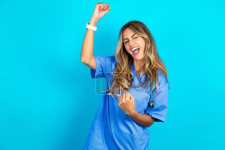 Téléchargez les photos : Photo de profil de excitée belle médecin femme debout sur fond bleu studio bonne humeur lever les poings hurlant réjouissant fan de basket-ball de sport - en image libre de droit