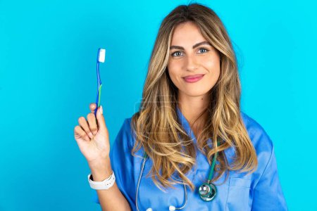 Foto de Hermosa doctora de pie sobre fondo azul estudio sosteniendo un cepillo de dientes y sonriendo. Concepto de salud dental. - Imagen libre de derechos