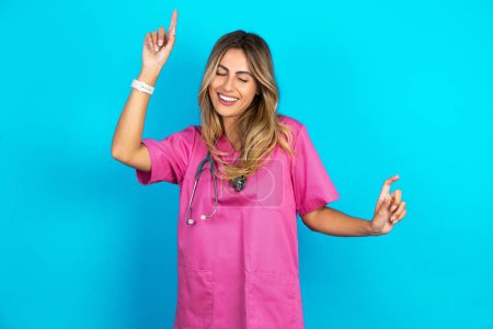 Téléchargez les photos : Photo de femme caucasienne optimiste médecin en uniforme médical rose avec stéthoscope s'amuse et danse vêtements insouciants être d'humeur parfaite fait des mouvements. Passe du temps libre sur disco partie - en image libre de droit