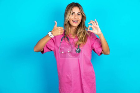 Téléchargez les photos : Femme caucasienne médecin en uniforme médical rose avec stéthoscope souriant et regardant heureux, insouciant et positif, geste de victoire ou de paix avec une main - en image libre de droit
