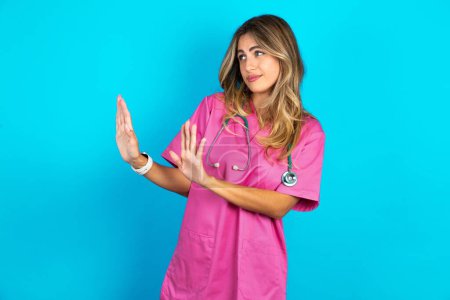Téléchargez les photos : Femme caucasienne mécontente médecin en uniforme médical rose avec stéthoscope garde les mains vers l'espace vide et demande de ne pas se rapprocher, voit quelque chose de désagréable - en image libre de droit