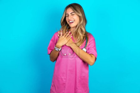 Foto de Médico mujer caucásica en uniforme médico rosa con estetoscopio expresa la felicidad, ríe agradablemente, mantiene las manos en el corazón - Imagen libre de derechos