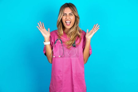 Foto de Crazy indignado médico caucásico mujer en uniforme médico rosa con estetoscopio grita en voz alta y gestos airadamente grita furiosamente. Emociones humanas negativas sentimientos concepto - Imagen libre de derechos