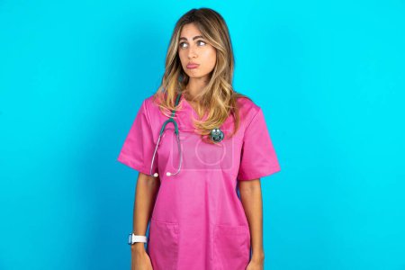 Photo for Dissatisfied beautiful doctor woman standing over blue studio background purses lips and has unhappy expression looks away stands offended. Depressed frustrated model. - Royalty Free Image