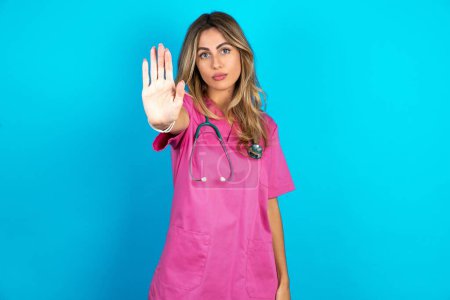 Photo for Caucasian woman doctor in pink medical uniform with stethoscope shows stop sign prohibition symbol keeps palm forward to camera with strict expression - Royalty Free Image