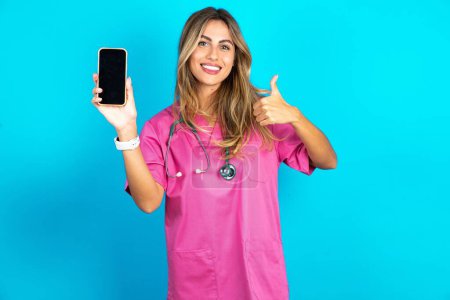 Photo for Caucasian woman doctor in pink medical uniform with stethoscope Show blank screen smartphone, thumb up recommend new app - Royalty Free Image