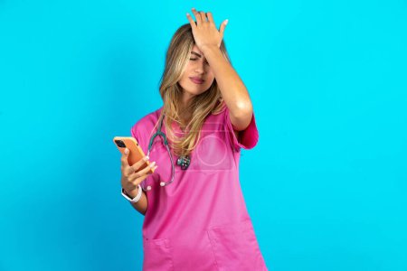 Photo for Upset depressed caucasian woman doctor in pink medical uniform with stethoscope makes face palm as forgot about something important holds mobile phone expresses sorrow and regret blames - Royalty Free Image