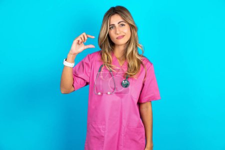 Foto de Mujer caucásica descontenta médico en uniforme médico rosa con formas de estetoscopio poco signo de mano demuestra algo no muy grande. Concepto de lenguaje corporal. - Imagen libre de derechos