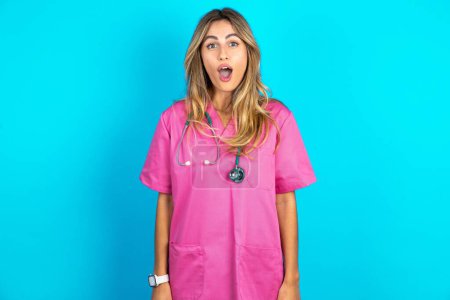 Téléchargez les photos : Choqué femme caucasienne médecin en uniforme médical rose avec stéthoscope fixe les yeux buggés garde la bouche ouverte a une expression surprise. Concept d'Omg - en image libre de droit