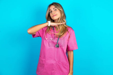 Photo for Caucasian woman doctor in pink medical uniform with stethoscope cutting throat with hand as knife, threaten aggression with furious violence. - Royalty Free Image