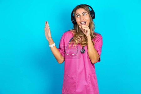 Foto de Mujer caucásica feliz médico en uniforme médico rosa con estetoscopio canta canción favorita mantiene la mano cerca de la boca como si el micrófono lleva auriculares inalámbricos, escucha música - Imagen libre de derechos