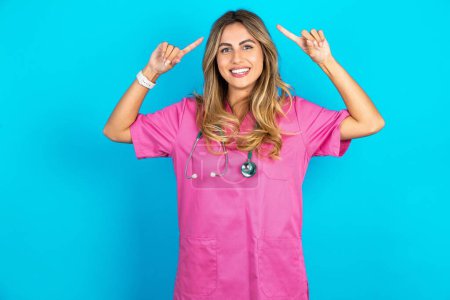 Foto de Foto de una loca doctora caucásica en uniforme médico rosa con estetoscopio apuntando con los dedos al cabello - Imagen libre de derechos