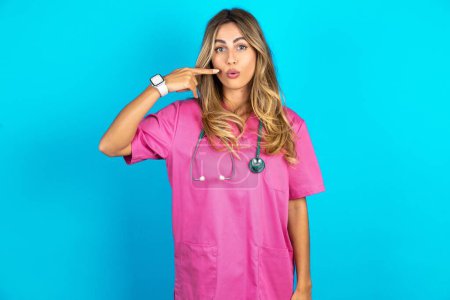 Foto de Encantadora doctora caucásica en uniforme médico rosa con estetoscopio que señala los labios de la puchera con el dedo índice, mostrando efecto después del procedimiento de elevación, - Imagen libre de derechos