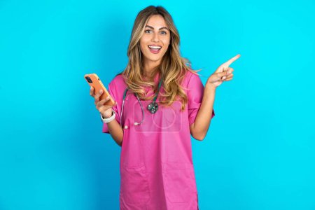 Téléchargez les photos : Femme caucasienne étonnante médecin en uniforme médical rose avec stéthoscope tenant son téléphone et pointant du doigt à côté de l'espace de copie vide - en image libre de droit