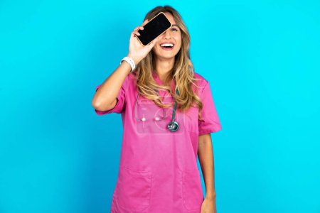 Foto de Médico mujer caucásica en uniforme médico rosa con estetoscopio sosteniendo teléfono inteligente moderno que cubre un ojo mientras sonríe - Imagen libre de derechos