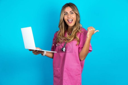 Téléchargez les photos : Femme caucasienne médecin en uniforme médical rose avec stéthoscope dirigeant espace vide tenir ordinateur portable - en image libre de droit