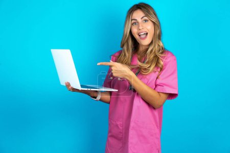 Photo for Shocked caucasian woman doctor in pink medical uniform with stethoscope pointing finger modern device - Royalty Free Image