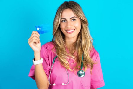 Photo for Photo of happy caucasian woman doctor in pink medical uniform with stethoscope recommend credit card - Royalty Free Image
