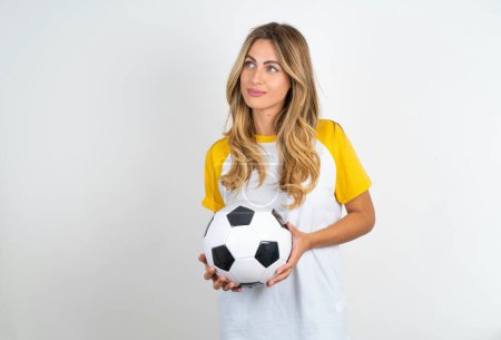 Photo for Charming thoughtful beautiful woman holding football ball over white background stands with arms folded concentrated somewhere with pensive expression thinks what to do - Royalty Free Image