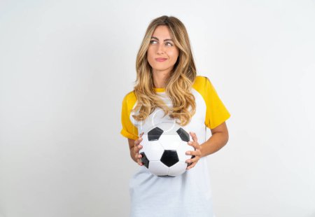 Photo for Beautiful woman holding football ball over white background steepled fingers and looks mysterious aside has great evil plan in mind - Royalty Free Image