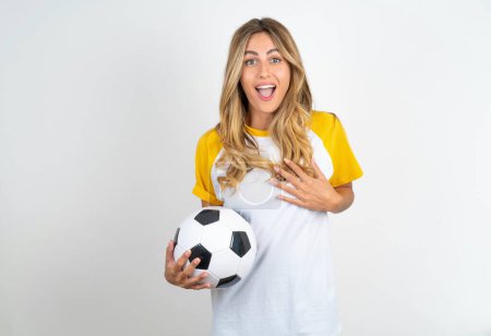 Foto de Hermosa mujer sosteniendo pelota de fútbol sobre fondo blanco sonrisas con dientes no puedo creer ojos expresa buenas emociones y sorpresa - Imagen libre de derechos