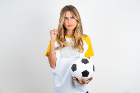 Photo for Irritated young beautiful woman holding football ball over white background blows cheeks with anger and raises clenched fists expresses rage and aggressive emotions. Furious model - Royalty Free Image