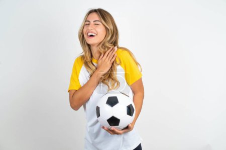Foto de Joven hermosa mujer sosteniendo pelota de fútbol sobre fondo blanco expresa felicidad, ríe agradablemente, mantiene las manos en el corazón - Imagen libre de derechos