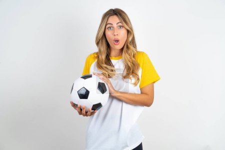 Photo for Surprised emotional young beautiful woman holding football ball over white background rubs palms and stares at camera with disbelief - Royalty Free Image