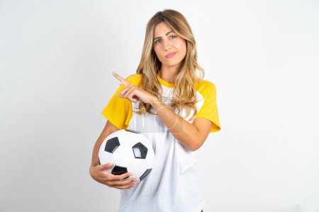 Photo for Serious young beautiful woman holding football ball over white background crosses hands and points at different sides hesitates between two items. Hard decision concept - Royalty Free Image