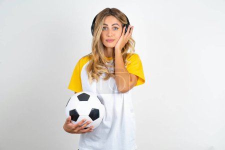 Photo for Serious displeased young beautiful woman holding football ball over white background  looks puzzled at camera being angry wears stereo headphones listens music while walking at street - Royalty Free Image