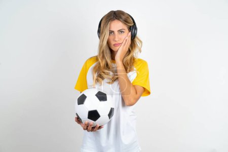 Foto de Retrato de triste joven hermosa mujer sosteniendo pelota de fútbol sobre fondo blanco con las manos en la cara - Imagen libre de derechos