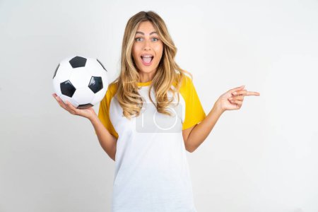 Foto de Mujer hermosa sorprendida vistiendo camiseta de fútbol sobre fondo blanco apuntando al espacio vacío sosteniendo pelota de fútbol - Imagen libre de derechos